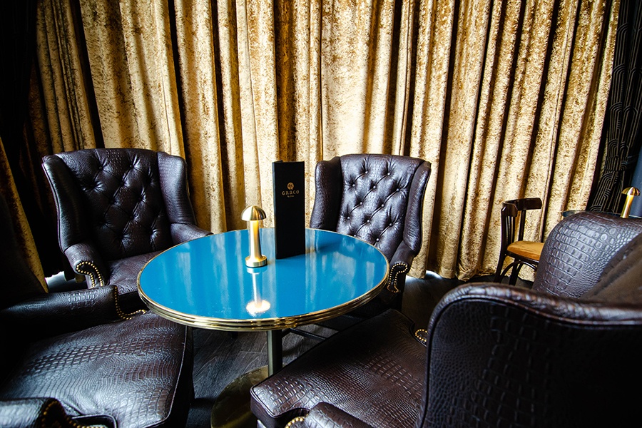Four brown leather armchairs surround a small teal table with heavy velour curtains behind.