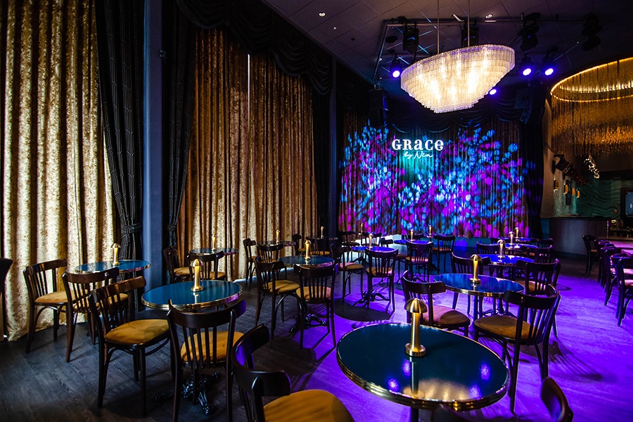 A dimly lit jazz club features heavy velour curtains, eye-catching chandeliers, and two-top tables, each with a small lamp.