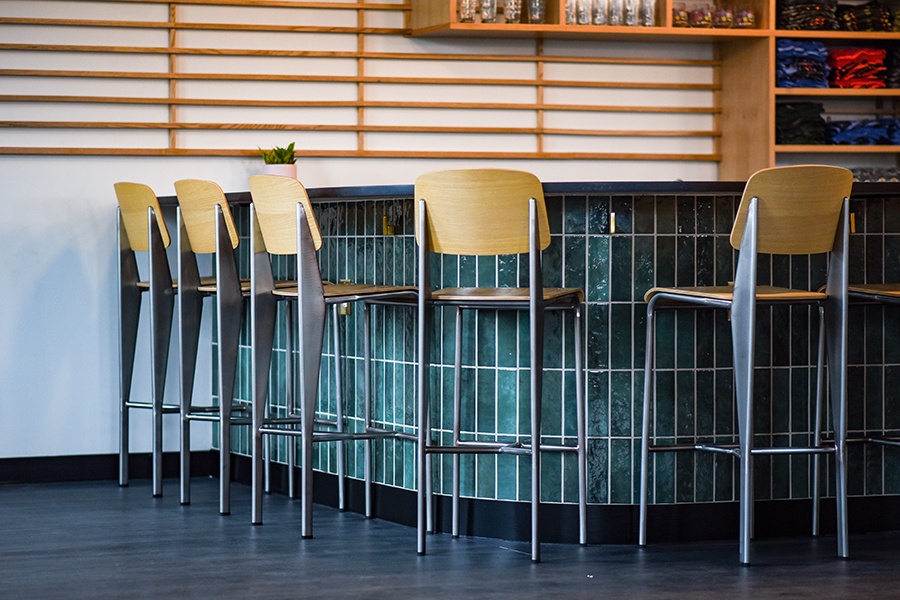 Light wooden chairs with steel frames line a green-tiled bar at a brewery taproom.
