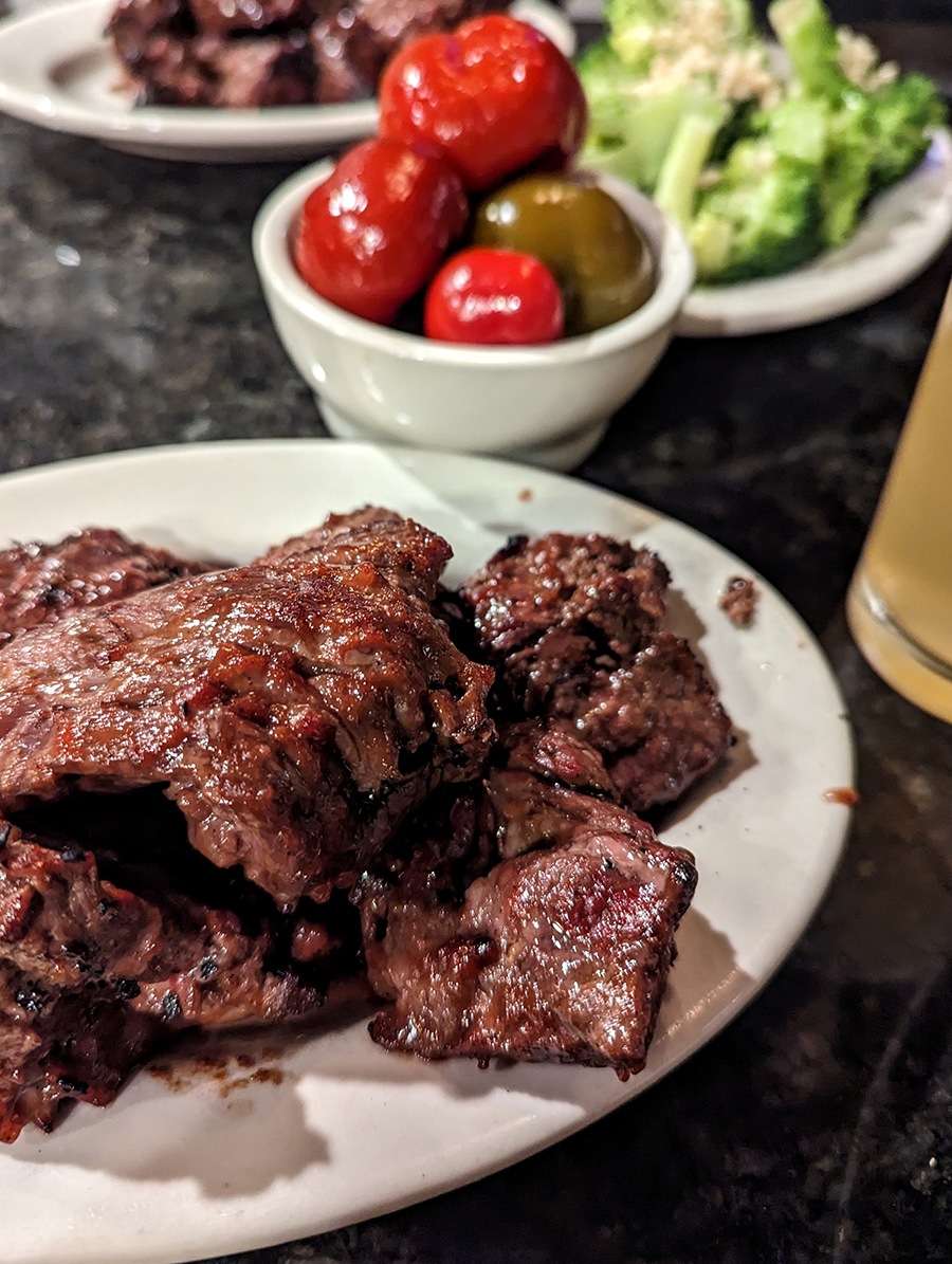 A plate is piled high with steak tips, with a small bowl of red and green cherry peppers and a plate of broccoli in the background.