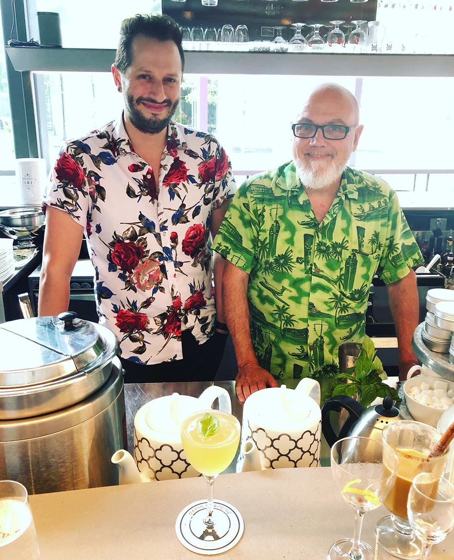 Two men stand smiling behind a bar, wearing Hawaiian shirts.