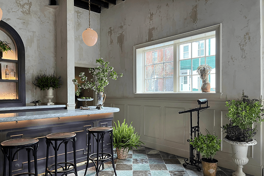 A sunny restaurant features a marble bar, lots of plants, a checkered floor, and light walls.