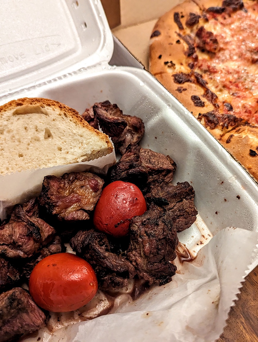 Steak tips are accompanied by two red cherry peppers and a thick slice of white bread in a styrofoam container. A cheese pizza is visible to the side.