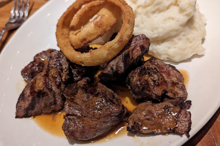 Steak tips are on a white plate, accompanied by onion rings and mashed potatoes.