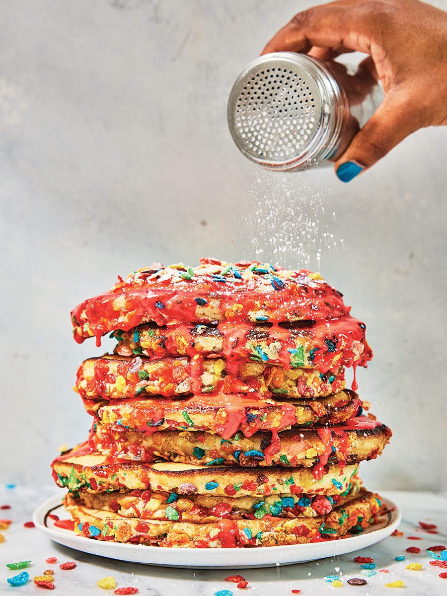A hand shakes powdered sugar onto a stack of eight fluffy pancakes, stuffed with Fruity Pebbles and drizzled with a pink cream.