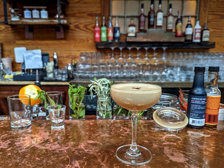 An espresso martini sits on a copper bar top with various glasses and liquor bottles in the background.