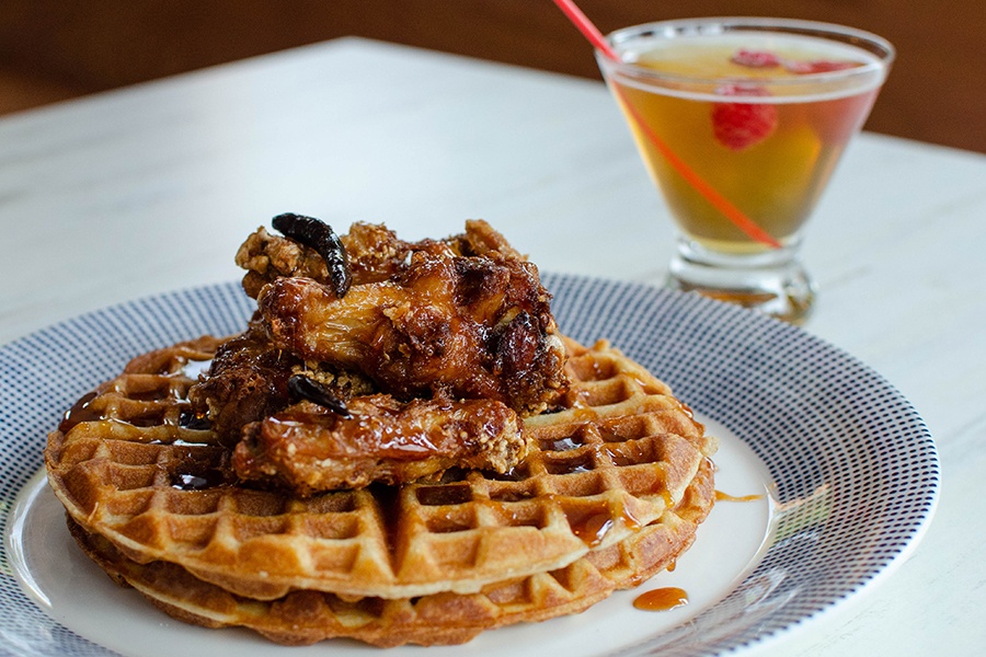 Crispy chicken wings sit atop two thin waffles, garnished with a Thai chili pepper and a drizzle of syrup. A pale brown cocktail with a raspberry in it is visible in the background.