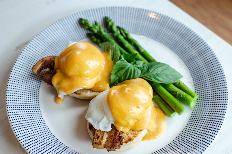 Thick pork belly and eggs sit atop an English muffin, smothered in a thick yellow sauce, with a side of asparagus.