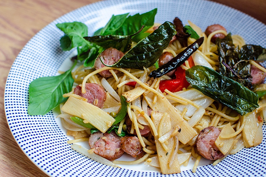 Spaghetti is mixed with slices of sausage, fresh basil, fried basil, bamboo shoots, and Thai chilis.