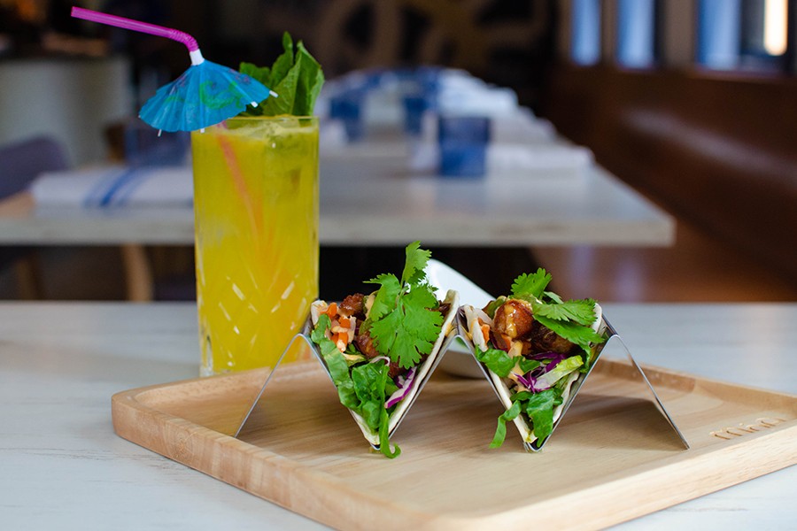 Two tacos are held upright in a metal holder with a yellow tropical cocktail to the side.