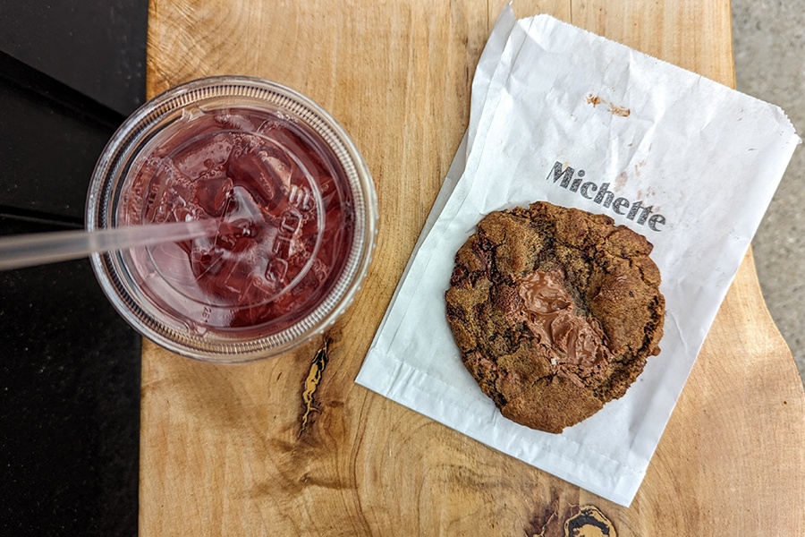 Overhead view of a chocolate chip cookie sitting on a paper bag that says Michette, next to a plastic cup of iced tea.