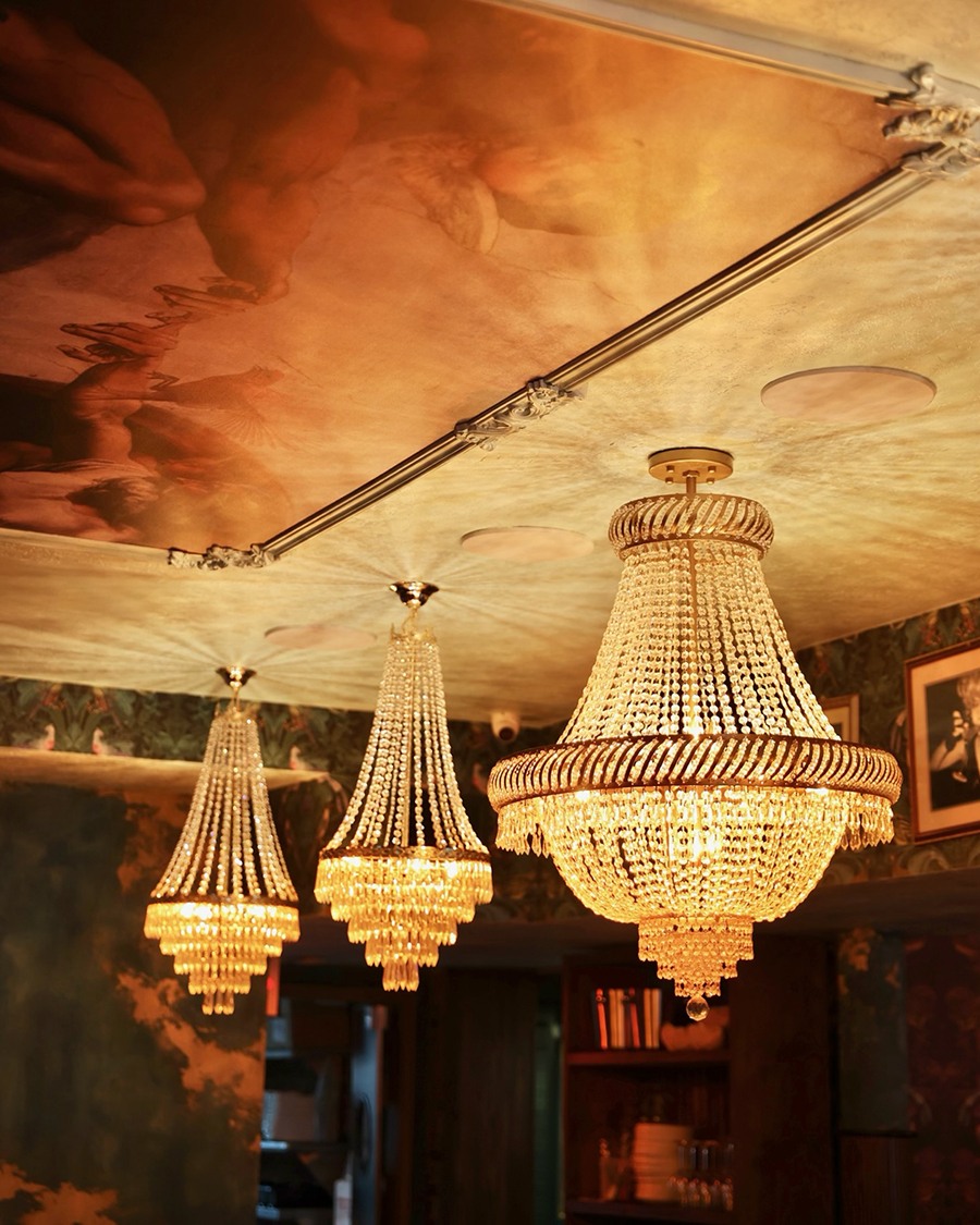 Three elegant chandeliers hang from a restaurant ceiling decorated with a Renaissance-style fresco.