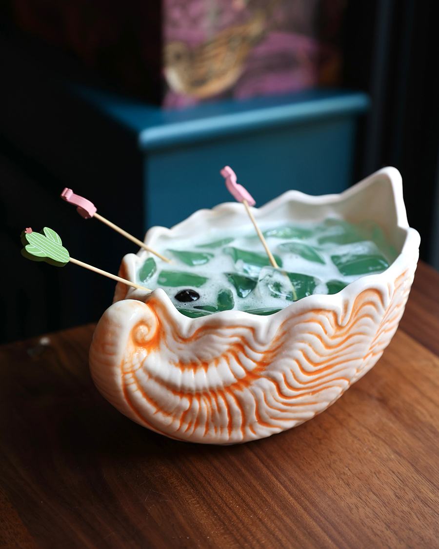 A big, blue cocktail is served in a conch shell cup, with maraschino cherries speared on flamingo and cactus toothpicks.