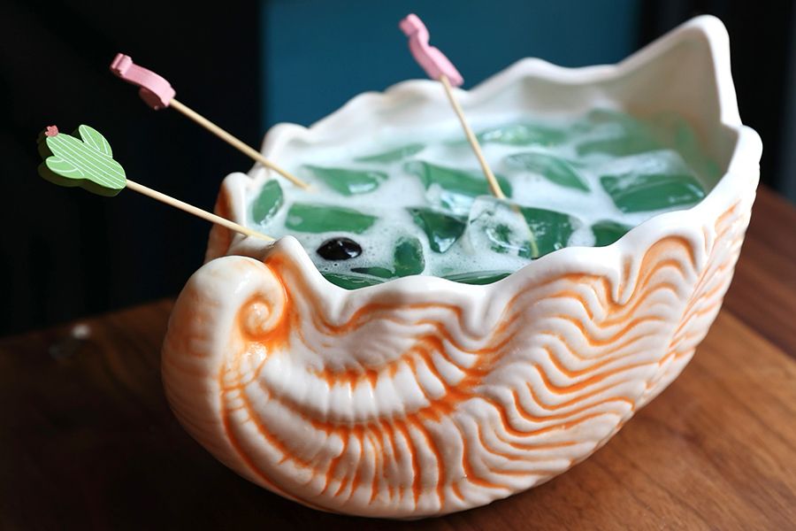 A big, blue cocktail is served in a conch shell cup, with maraschino cherries speared on flamingo and cactus toothpicks.