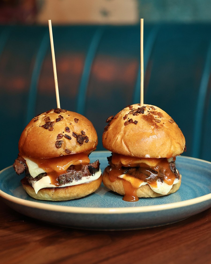 Two mini roast beef sandwiches sit on a teal plate on a wooden table, each speared with a wooden skewer.