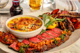 A rare steak is sliced and served with potatoes, creamed greens, a carafe of red wine, a lemon, and a bouquet of fresh herbs.