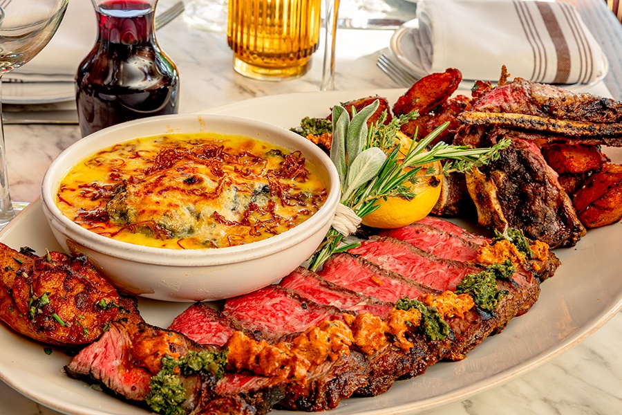 A rare steak is sliced and served with potatoes, creamed greens, a carafe of red wine, a lemon, and a bouquet of fresh herbs.