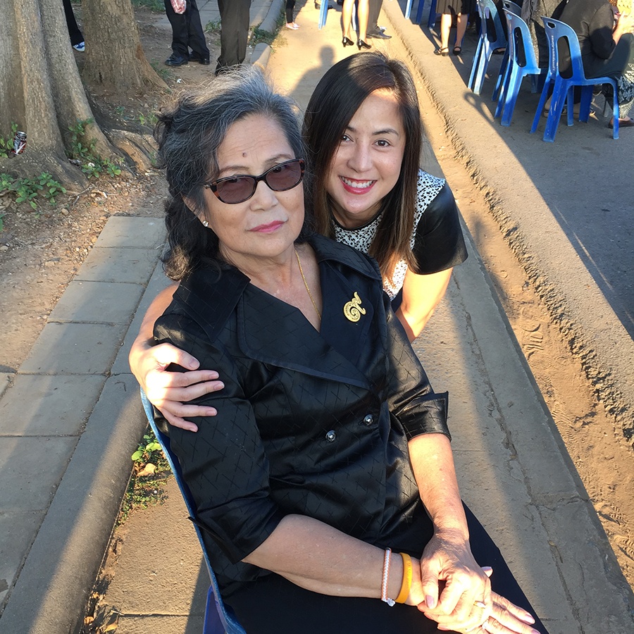 A mother and her adult daughter pose together, smiling at the camera, outdoors on a busy sidewalk.