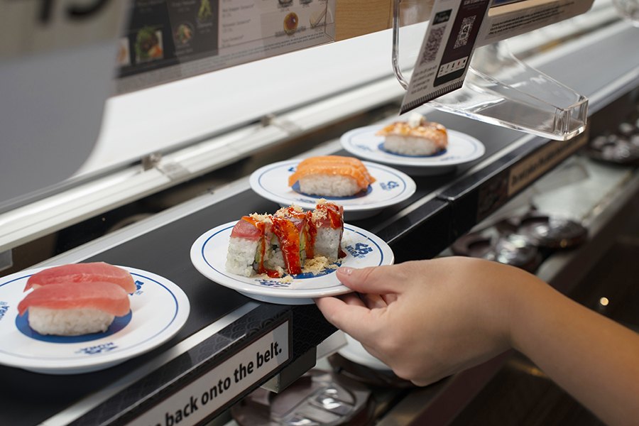 A hand grabs a plate with three pieces of sushi from a conveyor belt with other plates of different sushi.