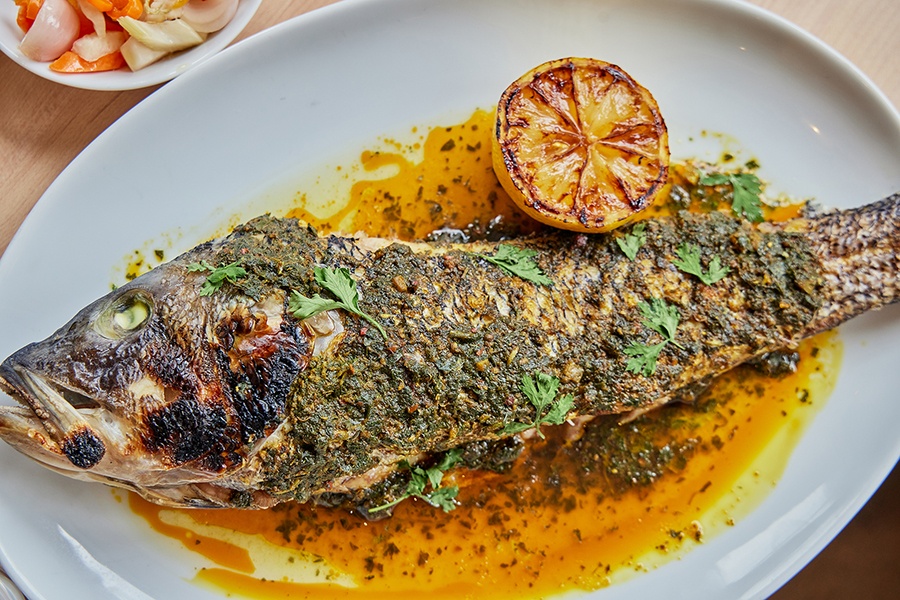 Overhead view of a whole roasted fish covered with a dark green herb-y pesto and parsley. A roasted half lemon sits to the side.