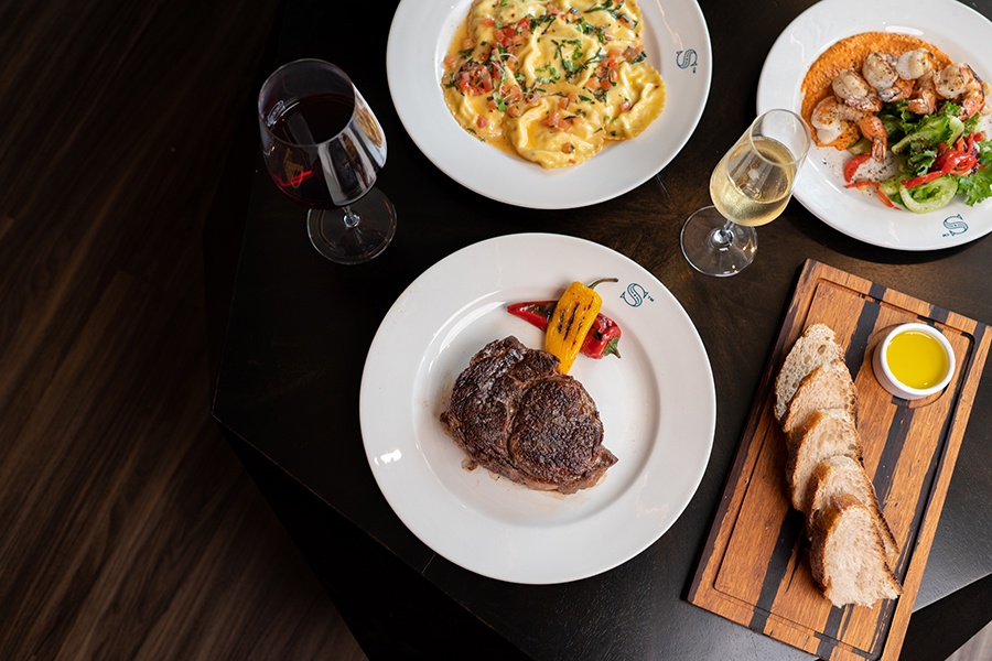 Overhead view of a dark wooden table with ravioli, grilled shrimp, steak, bread, and wine.