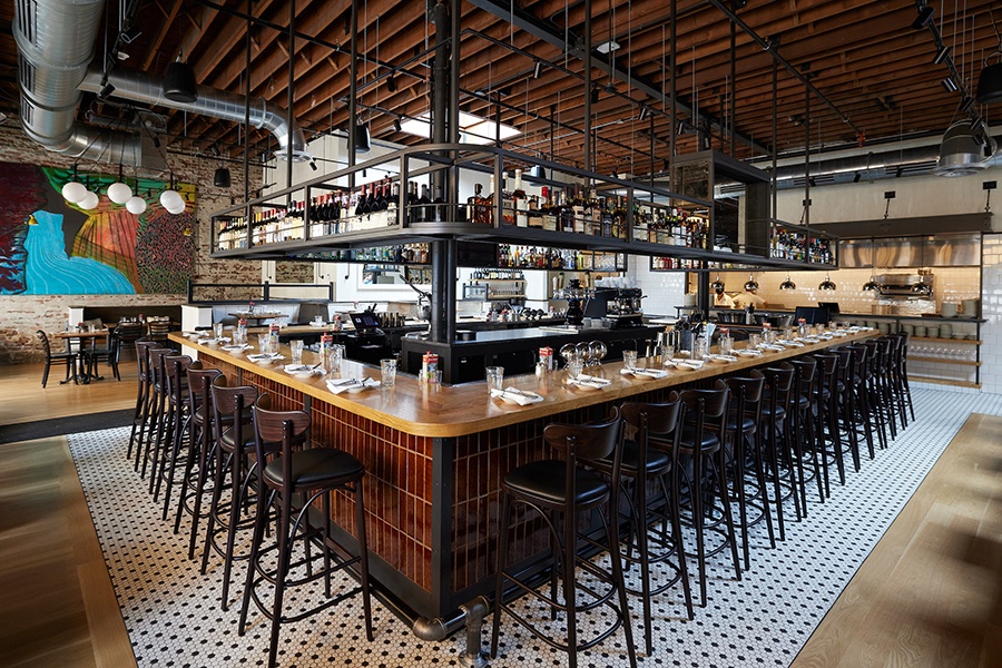 A large bar inside a breezy restaurant with high, metal-beam ceilings, black-and-white tiling, and a casual-but-not-too-casual feel.