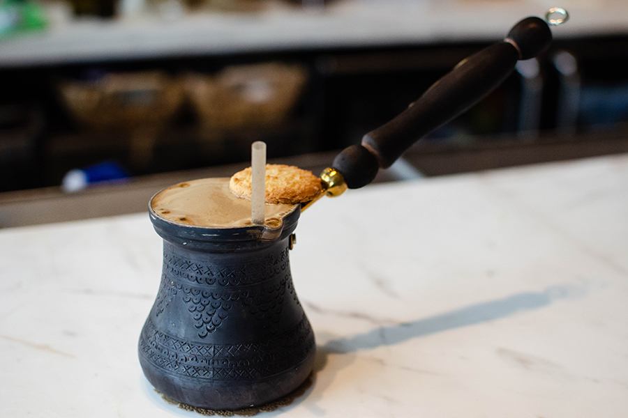 An espresso martini-like cocktail is served in a Greek briki and topped with a cookie.