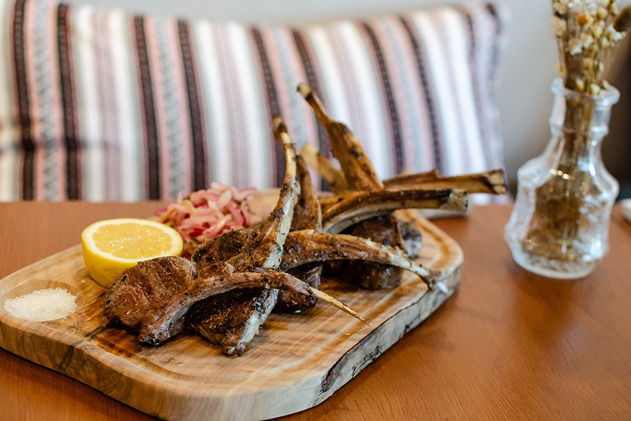 Lamb chops are served on a wooden platter with a half lemon, salt, and red onion.