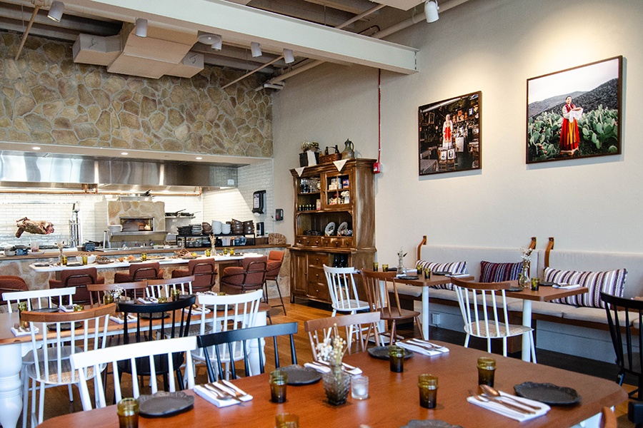 An restaurant interior has a Greek rustic feel, with large colorful photos of the Vlach people. A lamb on a rotisserie is visible in the open kitchen.