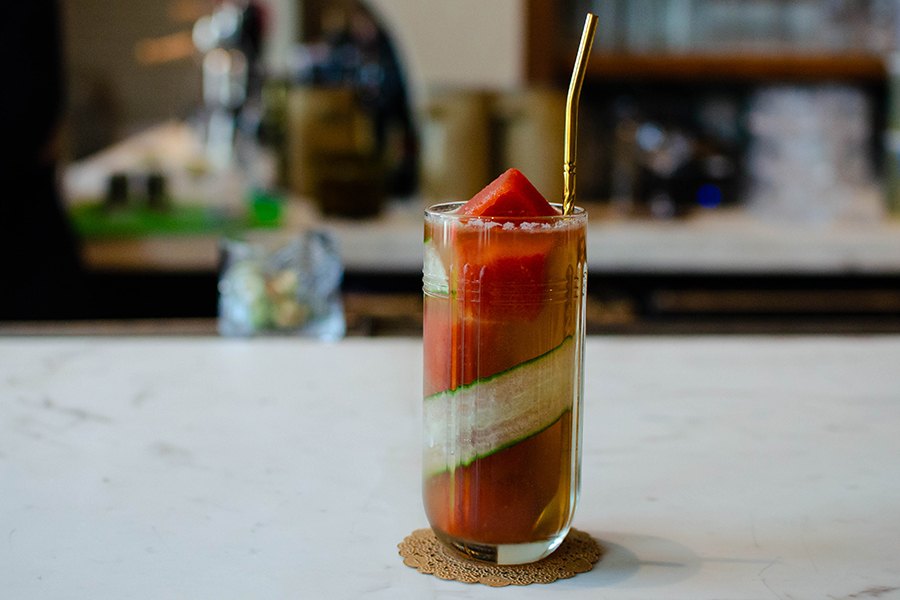 A tall cocktail is reddish-orange and has a ribbon of cucumber and red sorbet ice cubes.