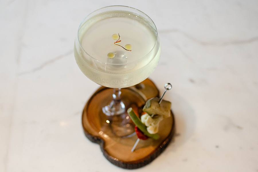 A dirty martini-style cocktail has drops of olive oil and a thread of saffron on top and a metal toothpick of pickled vegetables to the side.