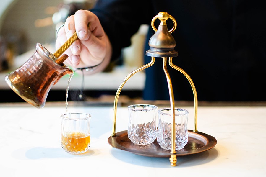 A hand pours an orange-brown liqueur into a delicate, small glass from a copper vessel.