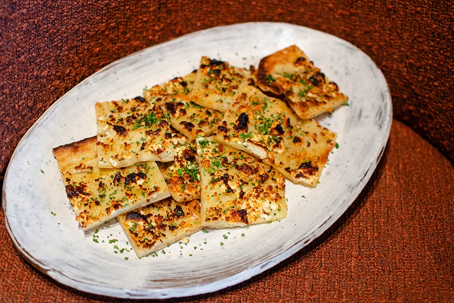 Thin rectangles of pita are topped with feta and herbs, browned in the oven, and piled on a rustic white plate on a maroon background.