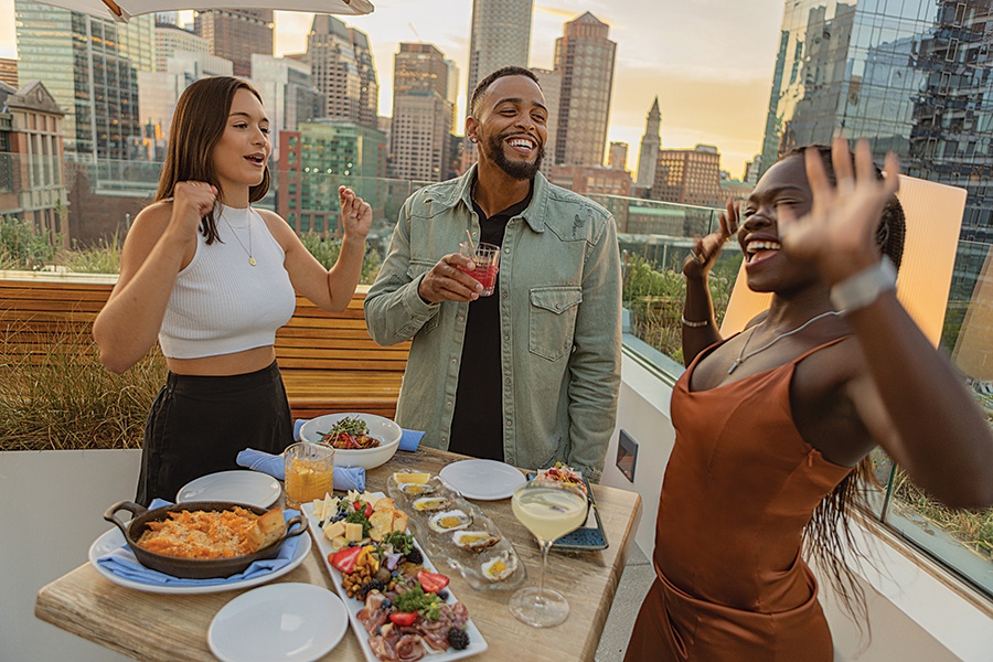 Happy people stand around a table full of food on a roof deck in the middle of a city.