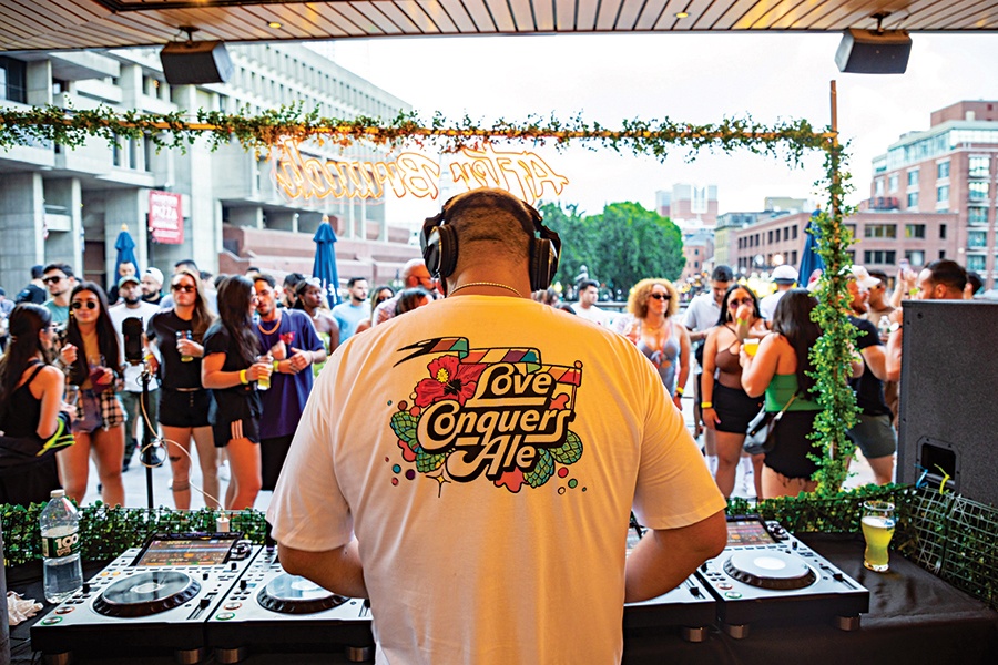 The back of a DJ is to the camera—his shirt says "Love Conquers Ale"—and people party in an outdoor space in front of him.