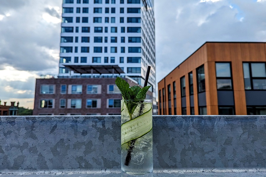 A clear mocktail, served tall with a slice of cucumber and sprig of mint, sits on a metal drink rail outdoors with city buildings in the background.