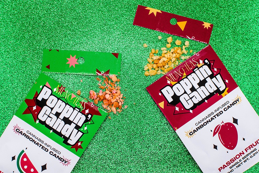 Two small packs of cannabis-infused pop rocks, one watermelon and one passion fruit, sit on a sparkly green background, open to reveal some of the pop rocks.
