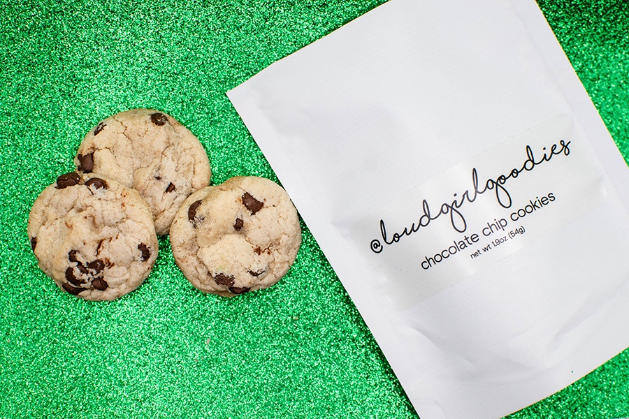 Three small chocolate chip cookies sit next to a white bag with loudgirlgoodies branding, all on a sparkly green background.