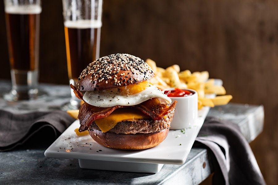A burger on a bun heavily seeded with black and white sesame is topped with melted orange cheese, bacon slices, and an egg. There's a side of fries with ketchup and beer visible in the background.
