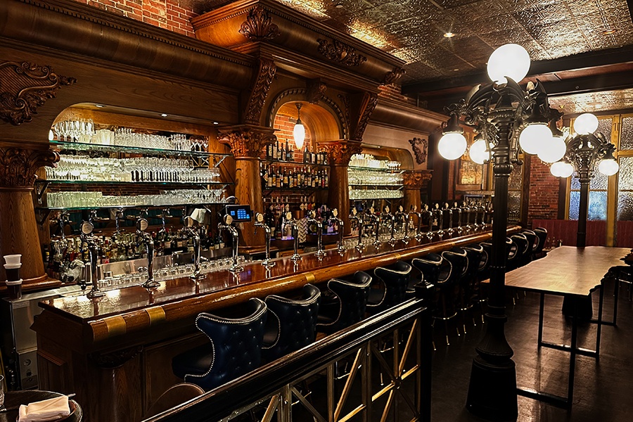A restaurant bar features a distinctive carved-wood backbar and lots of brick, black leather seats, and shiny wood.