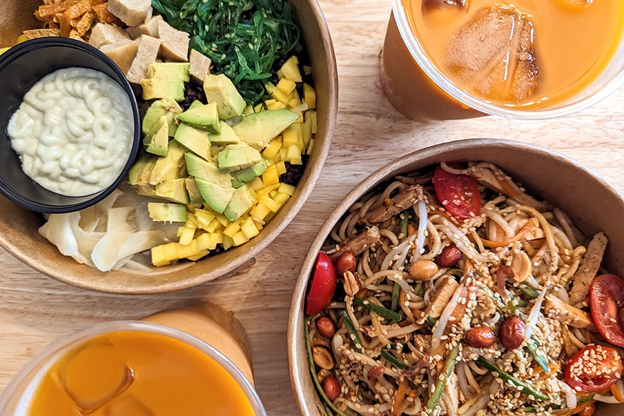 Overhead view of a rice bowl topped with avocado, seaweed, and mango, and a noodle bowl with peanuts, sesame seeds, and tomatos, with two bright orange Thai iced teas.
