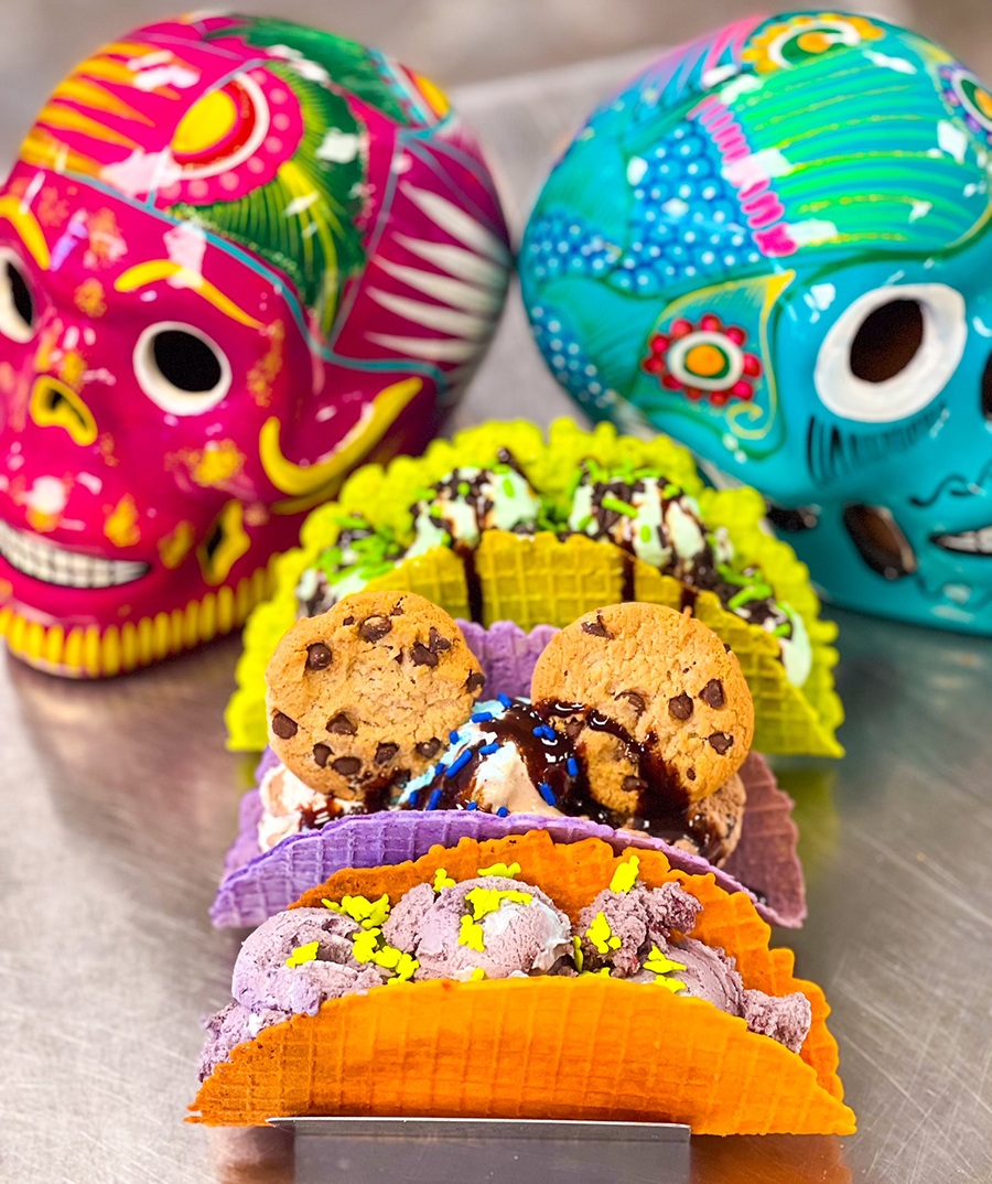 Three colorful waffle cones shaped like taco shells are stuffed with ice cream, cookies, and other toppings. Two colorful Mexican sugar skulls are in the background.