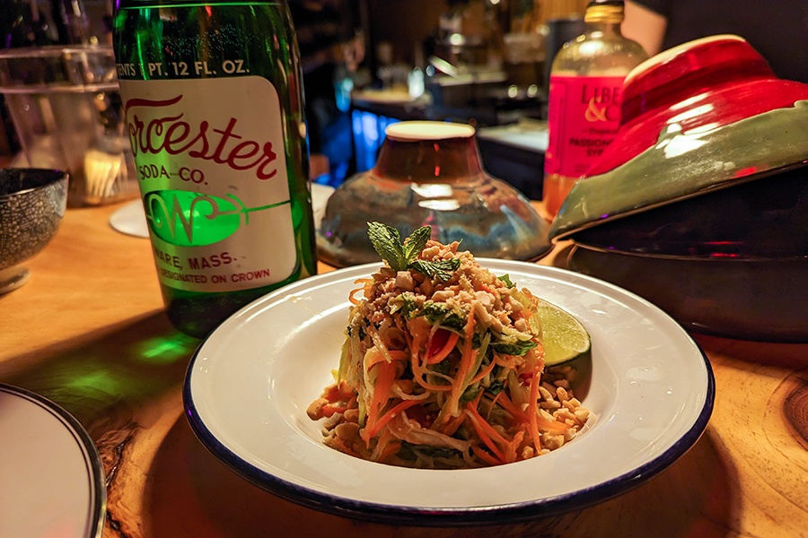A shredded mango salad with peanuts and a lime wedge is piled high on a white plate with a green glass bottle of water nearby.