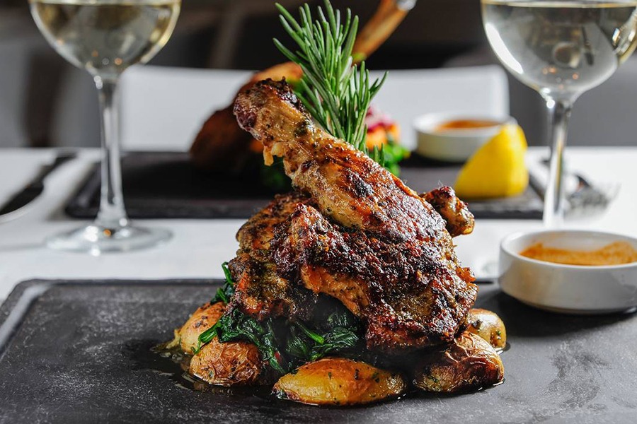 A roasted chicken sits atop a bed of potatoes with a sprig of rosemary coming up out of it.