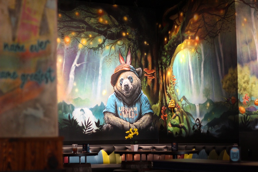 A large mural on a restaurant wall depicts a bear in a hat and a t-shirt that says urban wild, sitting in front of a colorful forest.