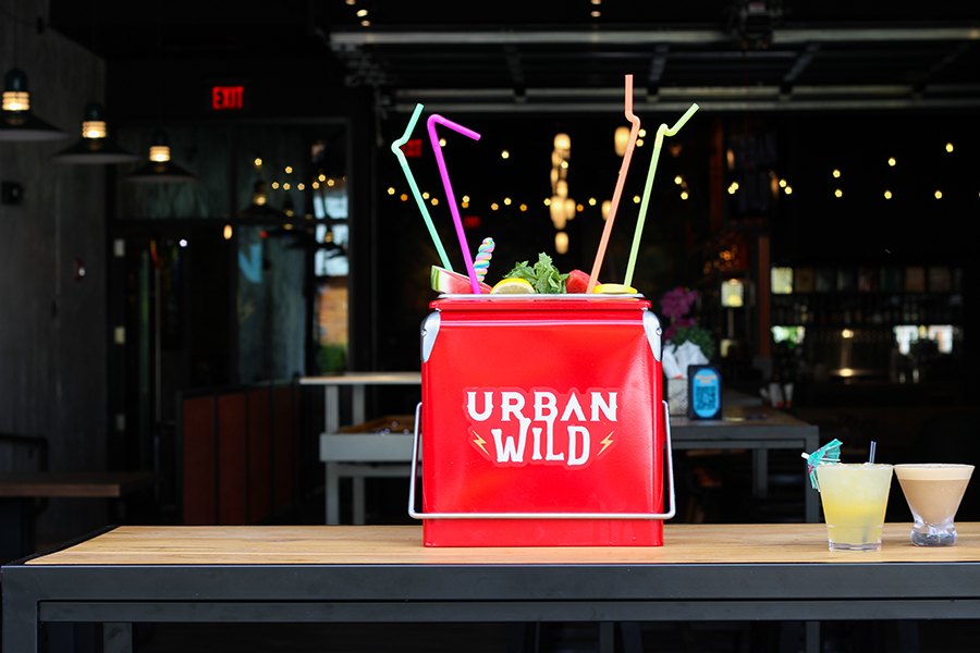 A bright red cooler that says Urban Wild is full of a drink topped with watermelon and lemon slices and colorful bendy straws.