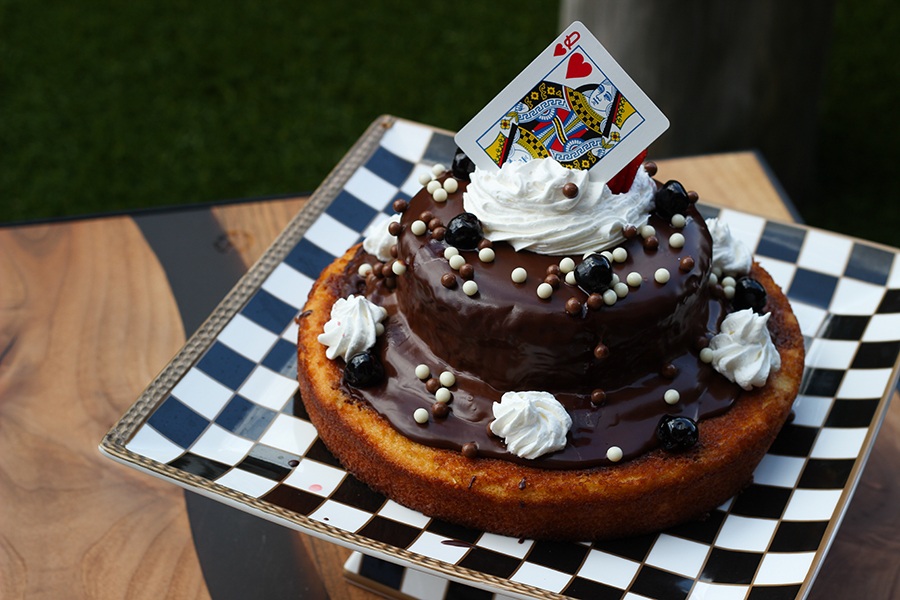 A two layer cake is covered with chocolate ganache, whipped cream, and a queen of hearts playing card, all served on a black and white checkered platter.