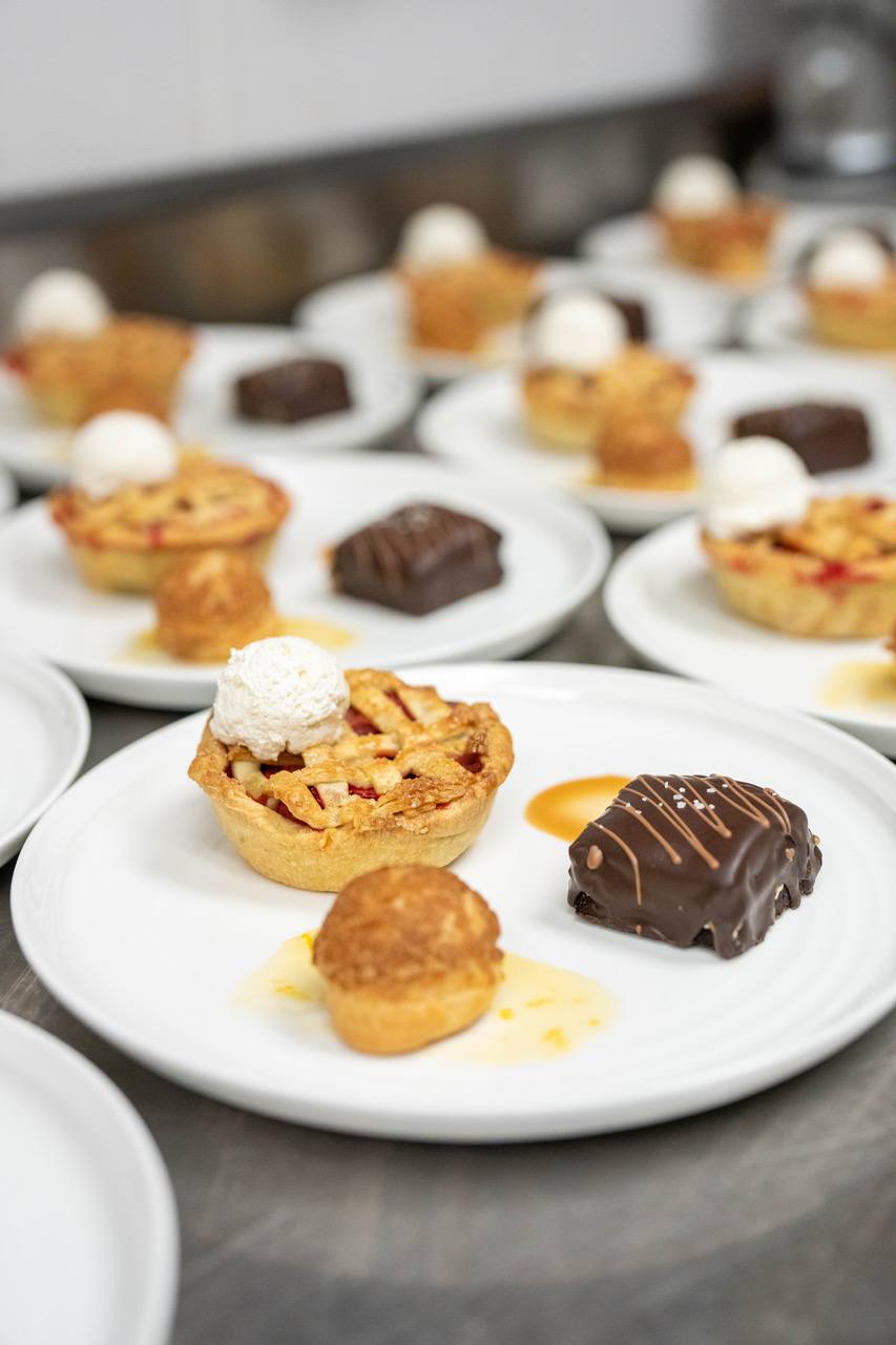 Many white plates are lined up, each with a small pie and two other miniature desserts.