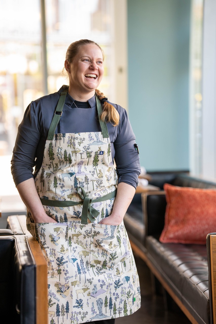 A woman in an apron stands, smiling, in a restaurant.