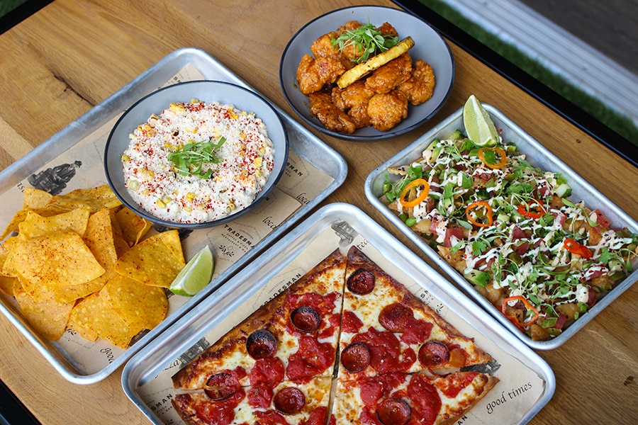Overhead view of a table full of bar snacks, including a thin-crust pizza, chips and dip, and more.